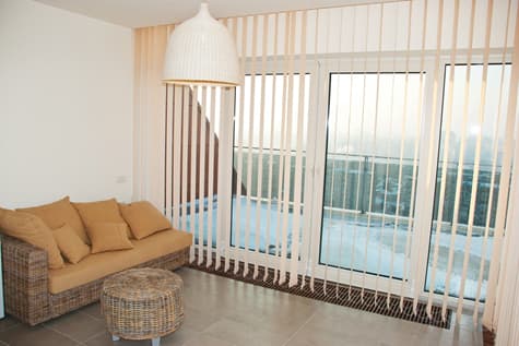 Leisure space featuring a thatch sofa and ottoman next to a large window with hanging vertical blinds.